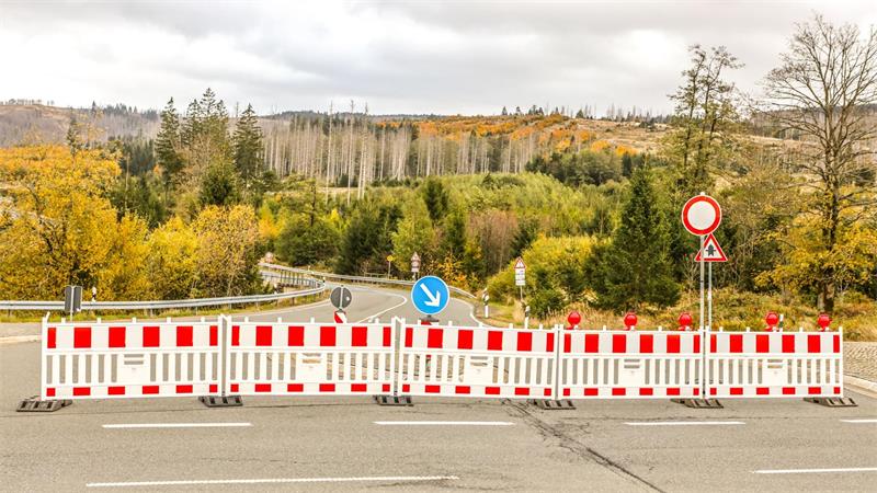 Absperrung mit rot-weißen Baken und mehreren Verkehrszeichen auf einer Straße vor herbstlicher Landschaft