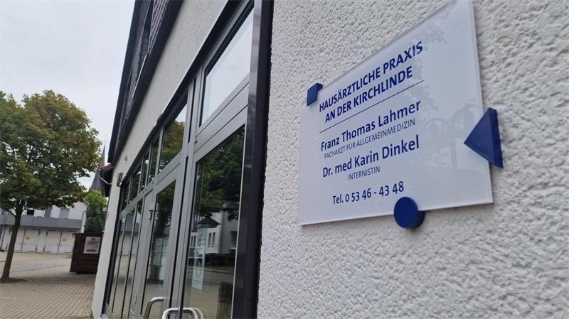 Schild an einer weißen Außenwand mit der Aufschrift 'Hausärztliche Praxis an der Kirchlinde' und den Namen zweier Ärzte neben einem modernen Gebäude mit Glasfassade.