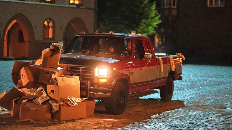 Roter Pickup auf Kopfsteinpflaster vor einem Haufen zerknitterter Kartons bei Nacht.
