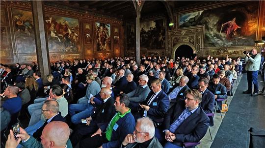 Großer Saal mit zahlreichen sitzenden Personen bei einer Veranstaltung vor kunstvoll verzierten Wänden und Decken
