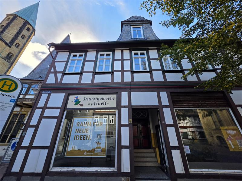 Fachwerkhaus mit Schaufenster und Schild 'Kunstgewerbe aktuell' sowie Plakat 'Raum für neue Ideen' neben einer Bushaltestelle und Kirchturm im Hintergrund