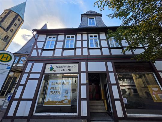 Fachwerkhaus mit Schaufenster und Schild 'Kunstgewerbe aktuell' sowie Plakat 'Raum für neue Ideen' neben einer Bushaltestelle und Kirchturm im Hintergrund