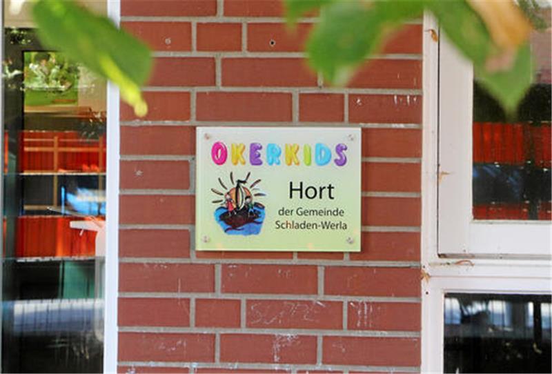 Schild mit bunter Aufschrift 'OKERKIDS Hort der Gemeinde Schladen-Werla' an roter Backsteinwand neben weißem Fensterrahmen