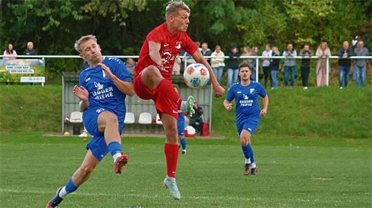 Zwei Fußballspieler in Aktion, einer in rotem Trikot springt und spielt den Ball, der andere in blauem Trikot versucht zu blocken, im Hintergrund Zuschauer am Spielfeldrand