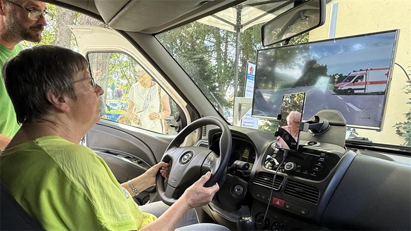 Eine Seniorin sitzt hinter dem Steuer eines Autos, vor der Frontscheibe ist ein Bildschirm mit einer Verkehrssituation zu sehen. 