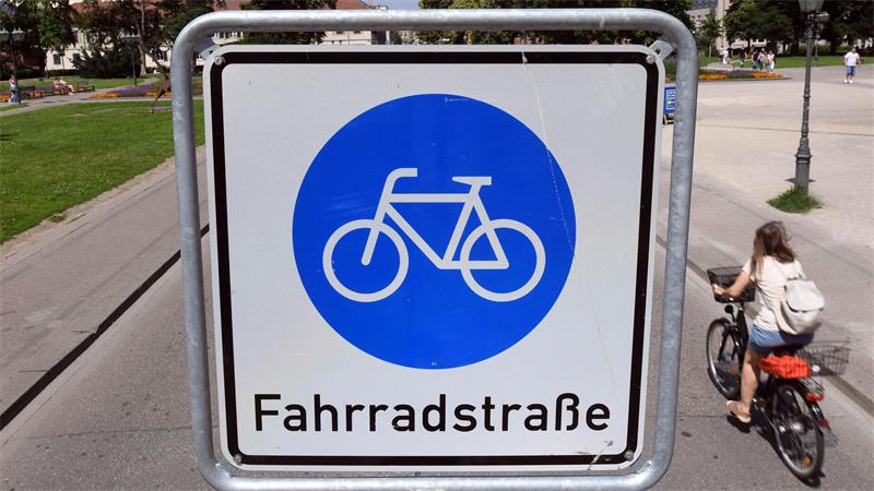 Verkehrsschild mit weißem Fahrradsymbol auf blauem Kreis und der Aufschrift 'Fahrradstraße' an einem Radweg, daneben fährt eine Frau mit Fahrrad.