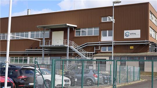 Mehrstöckiges Industriegebäude mit brauner Fassade, außenliegender Treppe und parkenden Autos hinter einem grünen Maschendrahtzaun