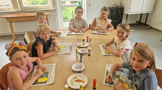 Sieben Kinder sitzen an einem Tisch und malen und basteln mit Papier und Klebestiften.