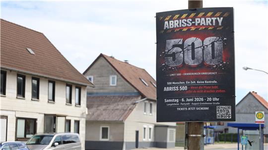 Straßenszene mit Wohnhäusern und einem großen Plakat für eine Abriss-Party mit der Zahl '501'