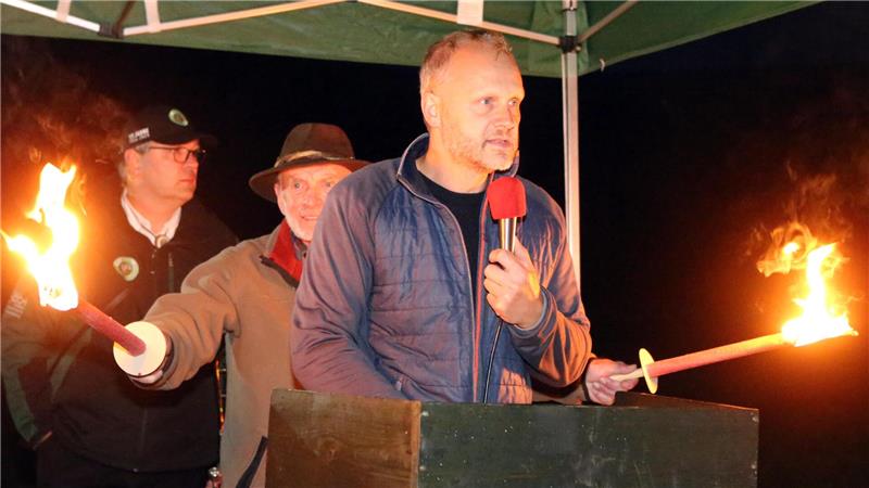 Alexander Ehrenberg hält eine Rede. Im Hintergrund steht ein Mann und sorgt mit Fackeln für Licht.