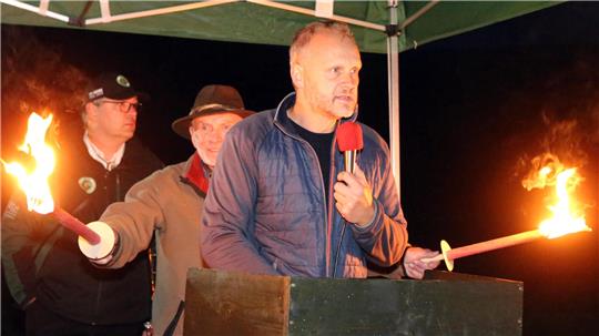 Alexander Ehrenberg hält eine Rede. Im Hintergrund steht ein Mann und sorgt mit Fackeln für Licht.