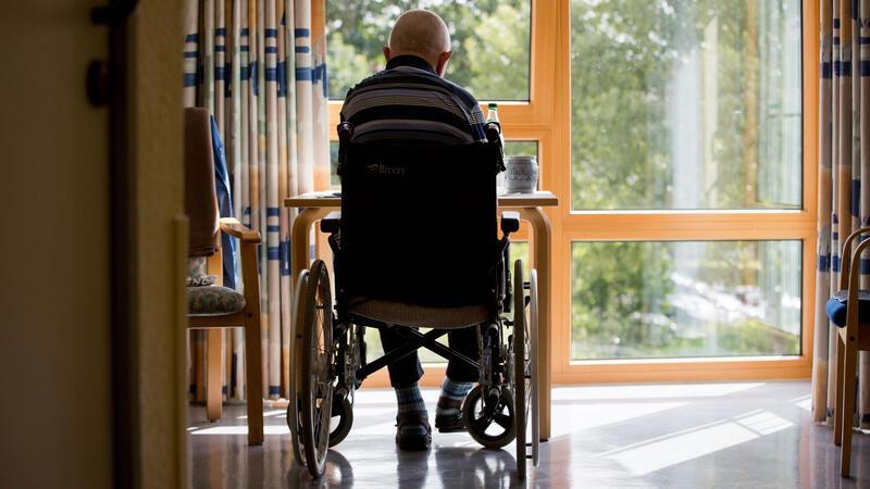 Person im Rollstuhl sitzt vor einem Fenster in einem hellen Raum mit Vorhängen und Tisch.