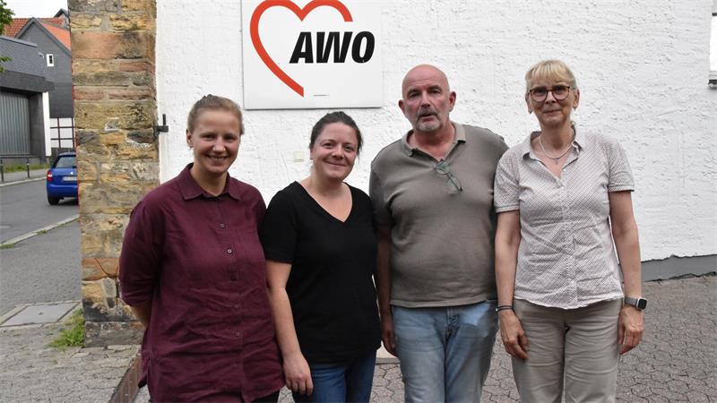 Vier Personen stehen vor einer weißen Wand mit einem roten Herz und dem Schriftzug 'AWO', zwei Frauen tragen kurzärmlige Hemden, ein Mann trägt ein graues Poloshirt.