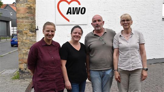 Vier Personen stehen vor einer weißen Wand mit einem roten Herz und dem Schriftzug 'AWO', zwei Frauen tragen kurzärmlige Hemden, ein Mann trägt ein graues Poloshirt.