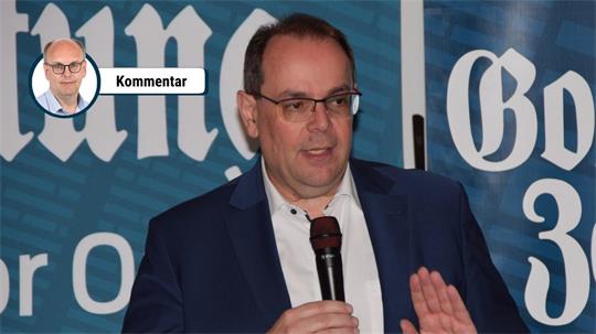 Mann mit Brille und dunklem Sakko hält Mikrofon und gestikuliert vor blauem Hintergrund mit weißer Schrift