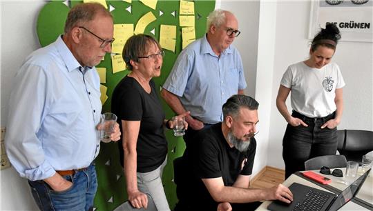 Vier Personen stehen und eine sitzt an einem Tisch mit Laptop in einem Raum mit grüner Wanddekoration und Post-its