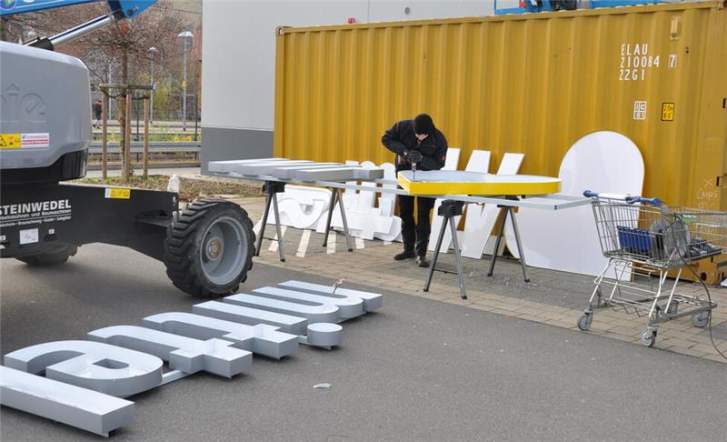Ein Mann in schwarzer Kleidung arbeitet an einem Edeka-Herz und großen weißen Buchstaben auf dem Boden neben einem gelben Container und einem blauen Hebegerät.
