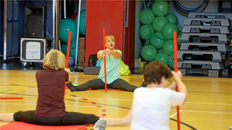 Drei Personen sitzen im Spagat auf dem Hallenboden und halten rote Stäbe vor sich, im Hintergrund grüne Gymnastikbälle und gestapelte Stepper.