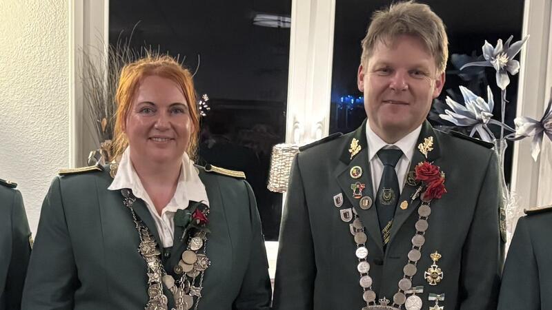 Zwei Personen in grünen Uniformjacken mit Medaillenketten und roten Ansteckblumen vor einer weißen Wand mit Fenster und Dekorationen