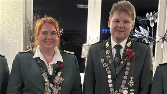 Zwei Personen in grünen Uniformjacken mit Medaillenketten und roten Ansteckblumen vor einer weißen Wand mit Fenster und Dekorationen
