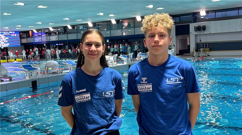 Zwei Personen in blauen LST-T-Shirts stehen an einem Hallenbad mit Schwimmern im Wasser und einer Anzeigetafel im Hintergrund