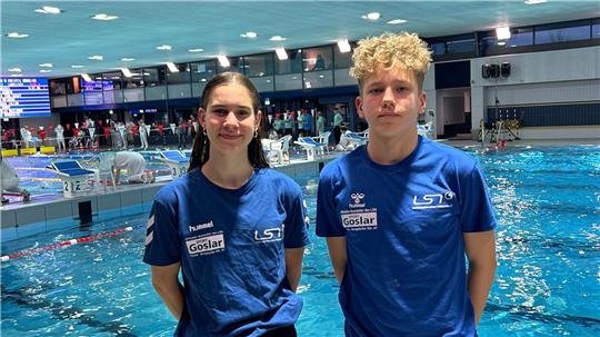 Zwei Personen in blauen LST-T-Shirts stehen an einem Hallenbad mit Schwimmern im Wasser und einer Anzeigetafel im Hintergrund
