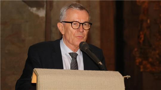 Älterer Mann mit grauen Haaren, Brille, dunklem Anzug, weißem Hemd und gemusterter Krawatte steht hinter einem Rednerpult mit Mikrofon in einem Innenraum