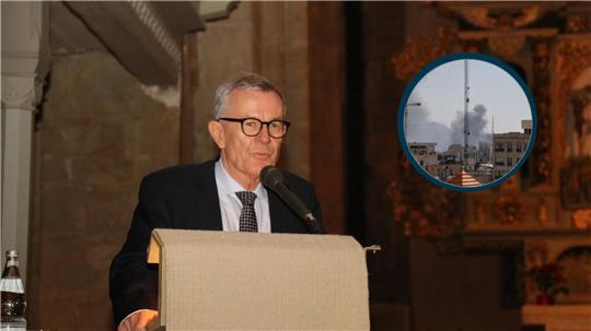 Älterer Mann mit grauen Haaren, Brille, dunklem Anzug, weißem Hemd und gemusterter Krawatte steht hinter einem Rednerpult mit Mikrofon in einem Innenraum