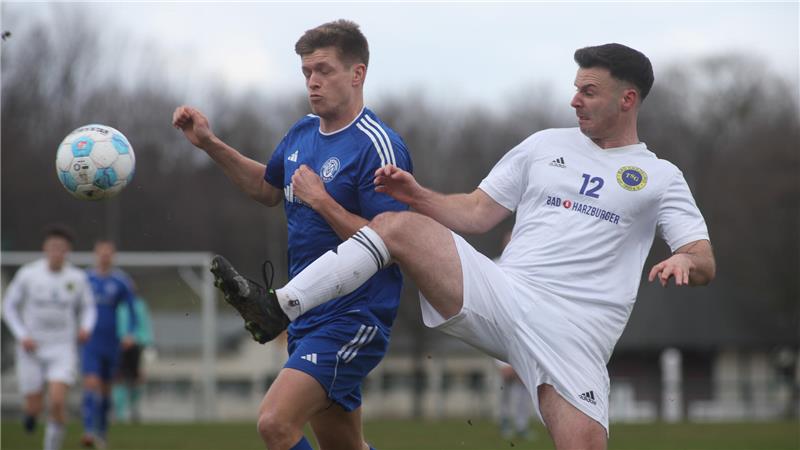 Zwei Fußballspieler in Aktion, einer in weißem Trikot mit Nummer 12 hebt das Bein zum Ball, der andere in blauem Trikot steht daneben