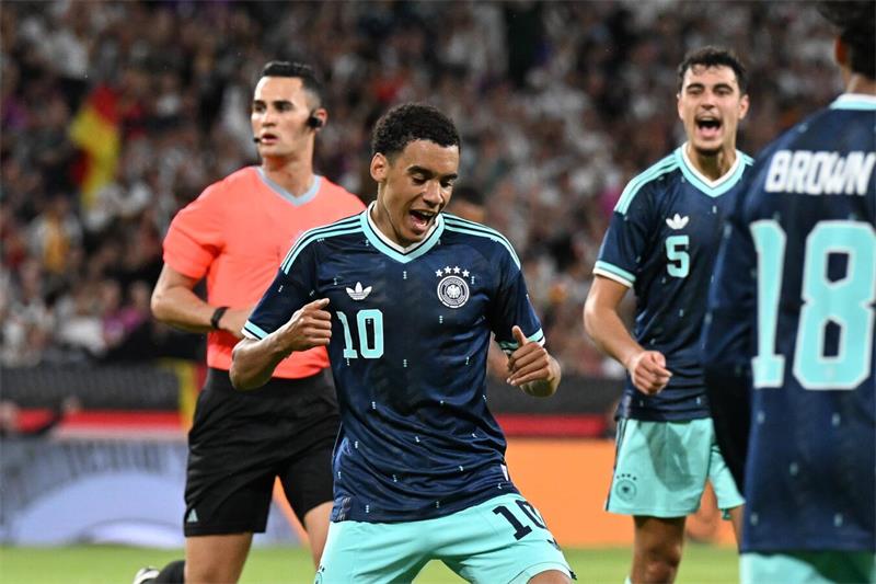 Ein Fußballer, der ein dunkleblaues Trikot und eine türkisfarbene Hose trägt jubelt mit beiden Armen über sein Tor. Links im Hintergrund ist ein Schiedsrichter, der ein rotes Trikot trägt. Rechts im Bild sind zwei weitere Fußballer in blauen Trikots zu sehen.