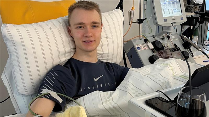 Person liegt in einem Krankenhausbett mit Infusionsschläuchen am Arm und medizinischen Geräten im Hintergrund.