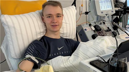 Person liegt in einem Krankenhausbett mit Infusionsschläuchen am Arm und medizinischen Geräten im Hintergrund.