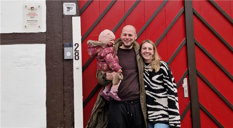 Zwei Erwachsene und ein Kind vor einem rot gestrichenen Holztor mit diagonal verlaufenden Latten, links ein weißes Haus mit braunen Holzbalken und einer Hausnummer 28.