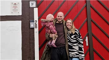 Zwei Erwachsene und ein Kind vor einem rot gestrichenen Holztor mit diagonal verlaufenden Latten, links ein weißes Haus mit braunen Holzbalken und einer Hausnummer 28.