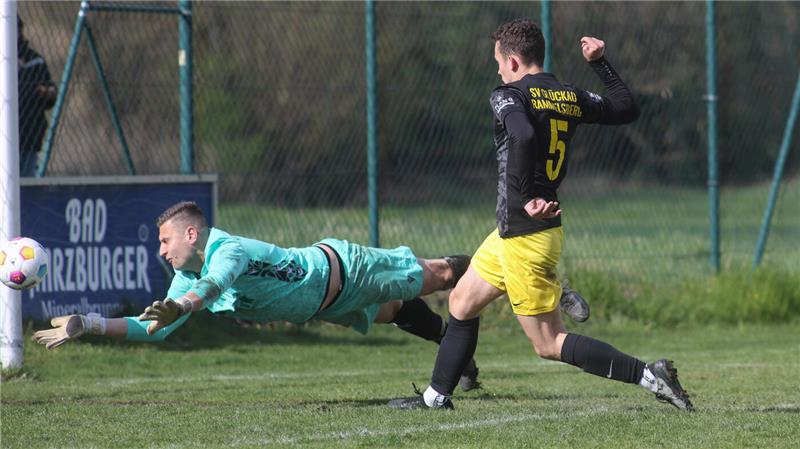 Torwart in türkisfarbenem Trikot taucht nach rechts, während ein Spieler in schwarzem Trikot und gelber Hose den Ball schießt