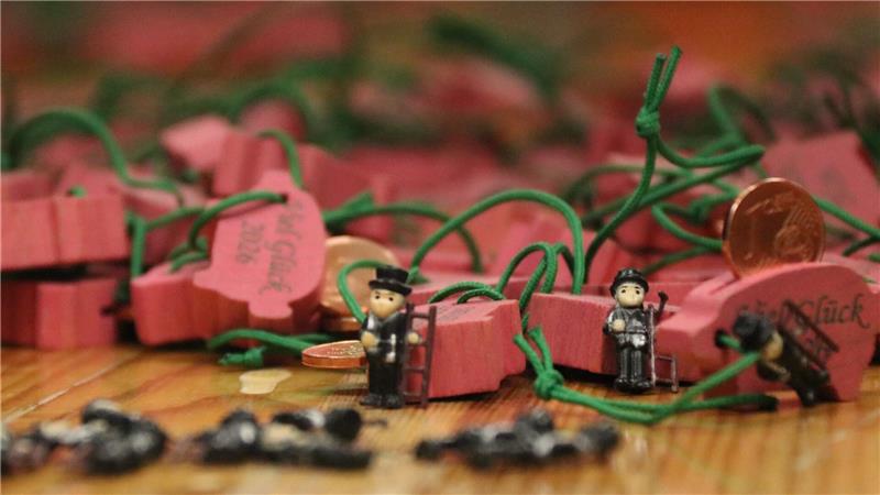 Der GZ-Schweinchenverkauf läuft in Goslar auf Hochtouren Mehrere rosa Holzschweinchen mit grünen Schnüren liegen auf einer Holzoberfläche, davor stehen zwei kleine Figuren mit schwarzen Hüten und grauer Kleidung