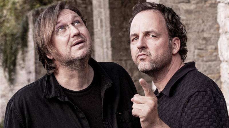 Zwei Männer, einer schaut nach oben, der andere zeigt auf die Kamera.