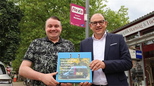 Zwei Männer stehen vor einem Baum und einem Geschäft, einer hält eine blaue Mappe mit einem Bild eines Lieferwagens und Text