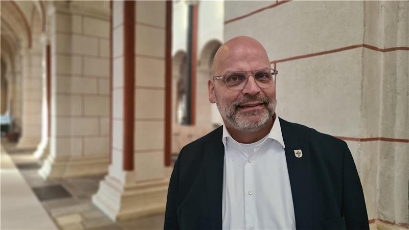 Ein Mann mit weißem Hemd und schwarzem Jacket steht im Seitenschiff einer Kirche und blickt freundlich in die Kamera. 