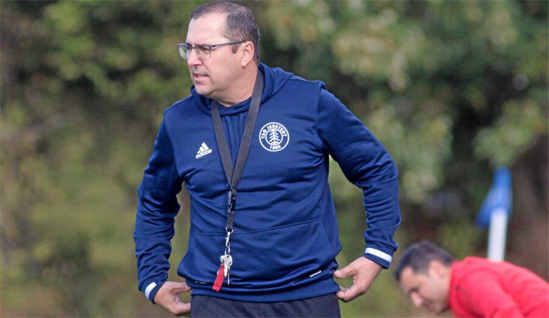 Mann mit Brille und blauem Trainingsanzug steht auf einem Sportplatz, im Hintergrund ein weiterer Mann in rotem Oberteil.