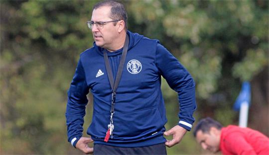 Mann mit Brille und blauem Trainingsanzug steht auf einem Sportplatz, im Hintergrund ein weiterer Mann in rotem Oberteil.