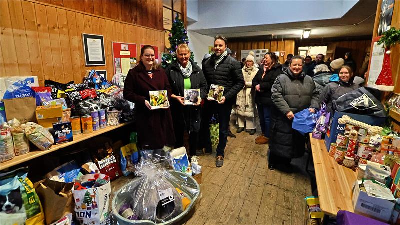 Drei Personen stehen vor einem Tisch mit zahlreichen Tierfutter- und Zubehörspenden, jede hält eine Tierkalender-Box, im Hintergrund ein geschmückter Weihnachtsbaum.