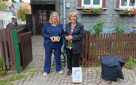 Zwei Frauen stehen vor einem Haus mit Schieferfassade und Blumenkästen an den Fenstern eine Frau hält eine Rose und vor ihnen steht ein Porträtfoto auf dem Boden.