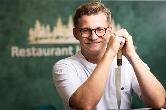 Ein Mann lächelt in die Kamera, in der Hand hält er ein Küchenmesser.