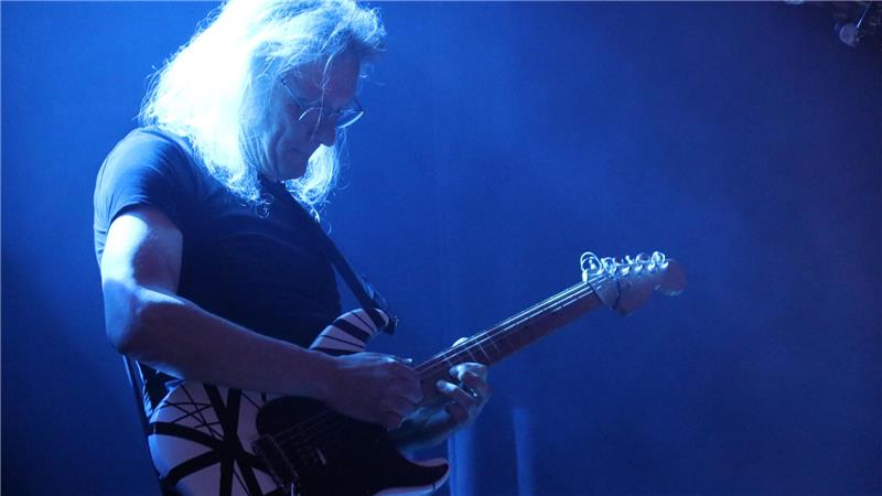 Musiker mit langen Haaren spielt E-Gitarre auf Bühne mit blauem Bühnenlicht.