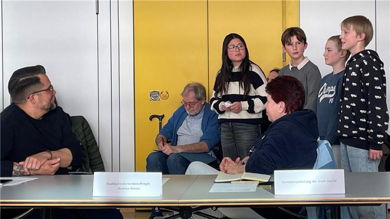 Gruppe von Menschen in einem Raum mit gelber Wand, einige sitzen an einem Tisch mit Namensschildern, andere stehen davor.