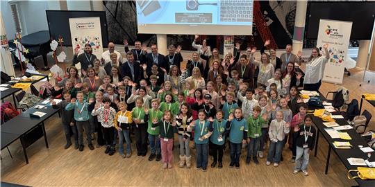 Große Gruppe von Kindern und Erwachsenen in einem Saal, viele Kinder tragen grüne oder blaue Pullover und halten kleine Objekte in den Händen