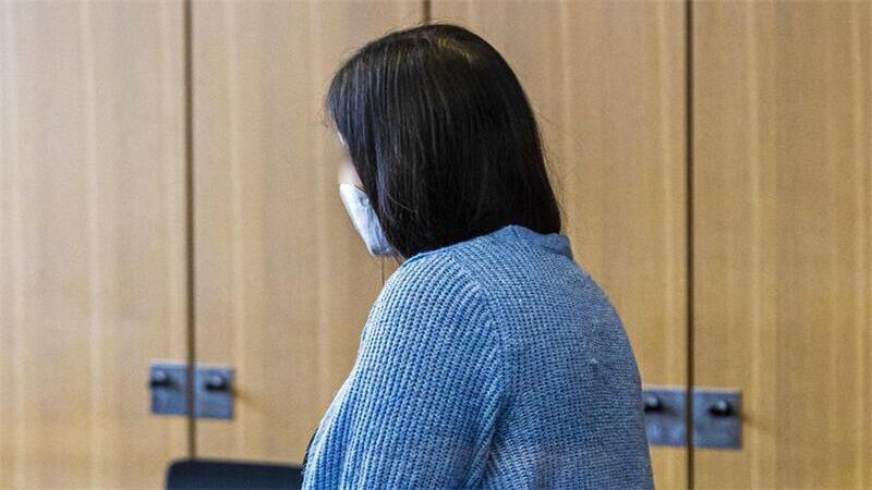 Frau mit dunklen Haaren und blauem Strickpullover steht seitlich vor einer Holzwand mit schwarzen Stühlen im Hintergrund