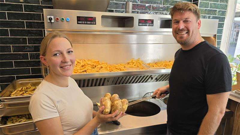 Zwei lächelnde Personen stehen vor einer Fritteuse, die mit Pommes gefüllt ist, eine Person hält mehrere Kartoffeln in den Händen