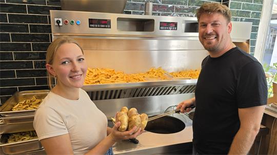 Zwei lächelnde Personen stehen vor einer Fritteuse, die mit Pommes gefüllt ist, eine Person hält mehrere Kartoffeln in den Händen